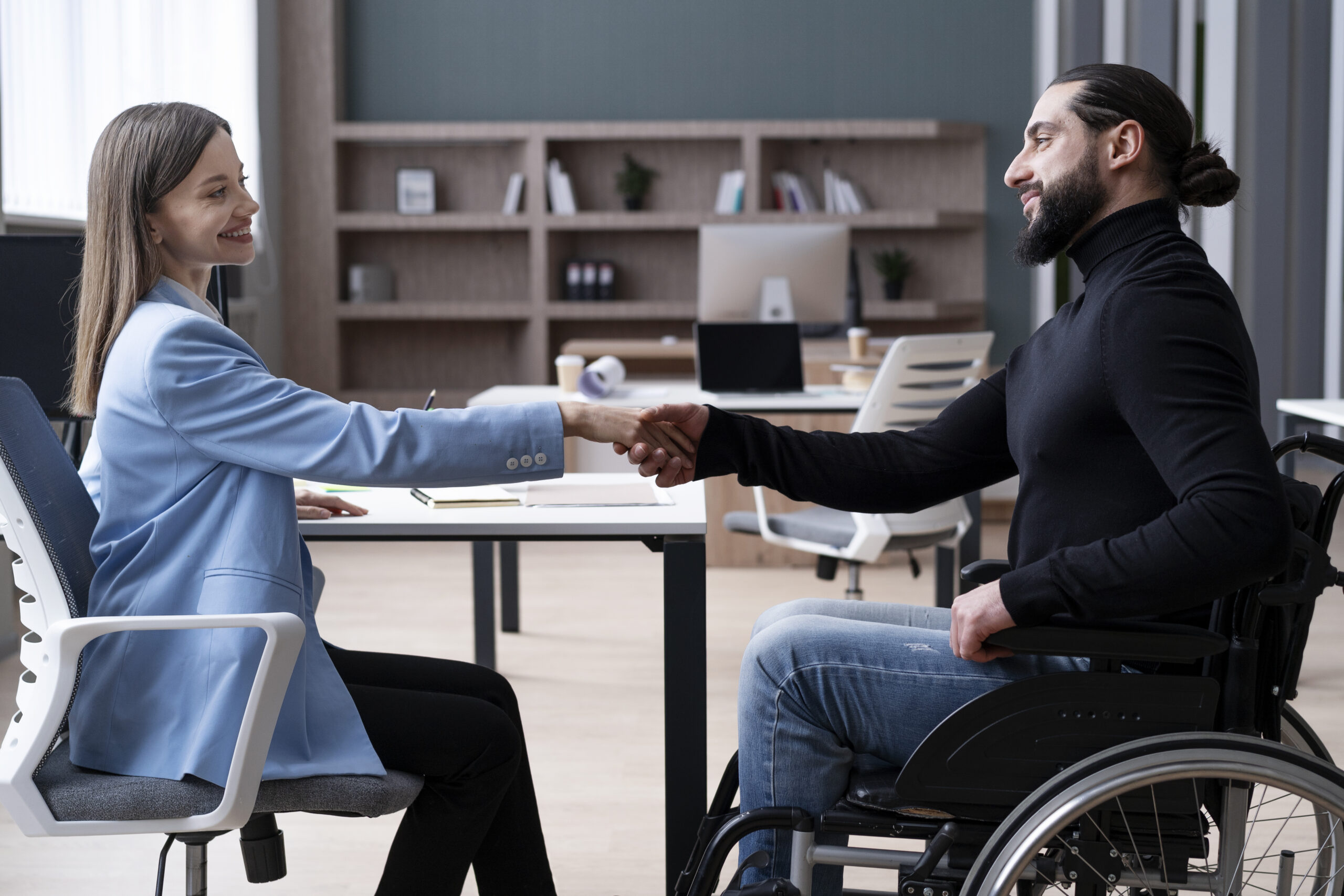 Two individuals shaking hands across a table in an office environment, one seated in a wheelchair.